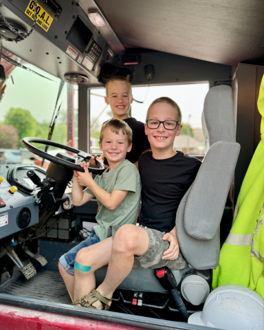 An image of three children sitting inside a firetruck at the steering wheel; all three children are smiling and posing for their photo being taken.